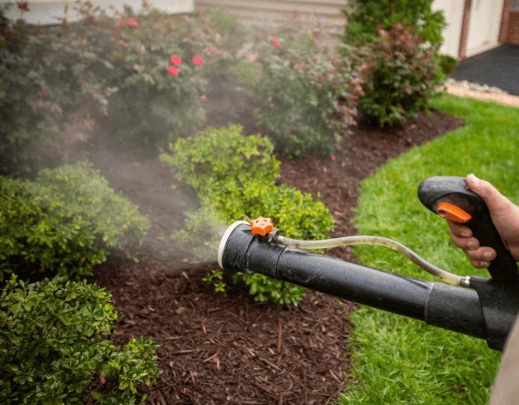 Technician’s hand holding a spraying wand dispersing mist along a home’s foundation and shrub line to control mosquitoes.