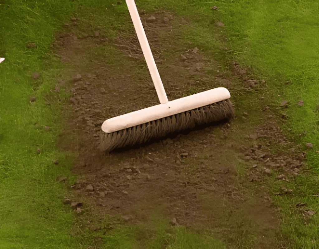 Close view of a push broom smoothing dark top-dressing soil into patches on a green lawn, finalizing the leveling and soil enrichment process.