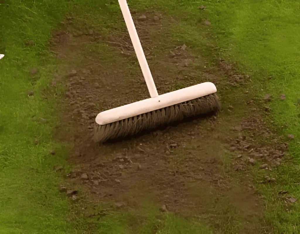 Close view of a push broom smoothing dark top-dressing soil into patches on a green lawn, finalizing the leveling and soil enrichment process.