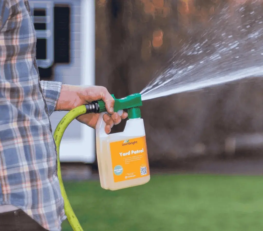 Close-up of a technician’s hand operating a hose-end sprayer dispersing mosquito control solution over a lawn and landscape, creating an insect-free barrier.