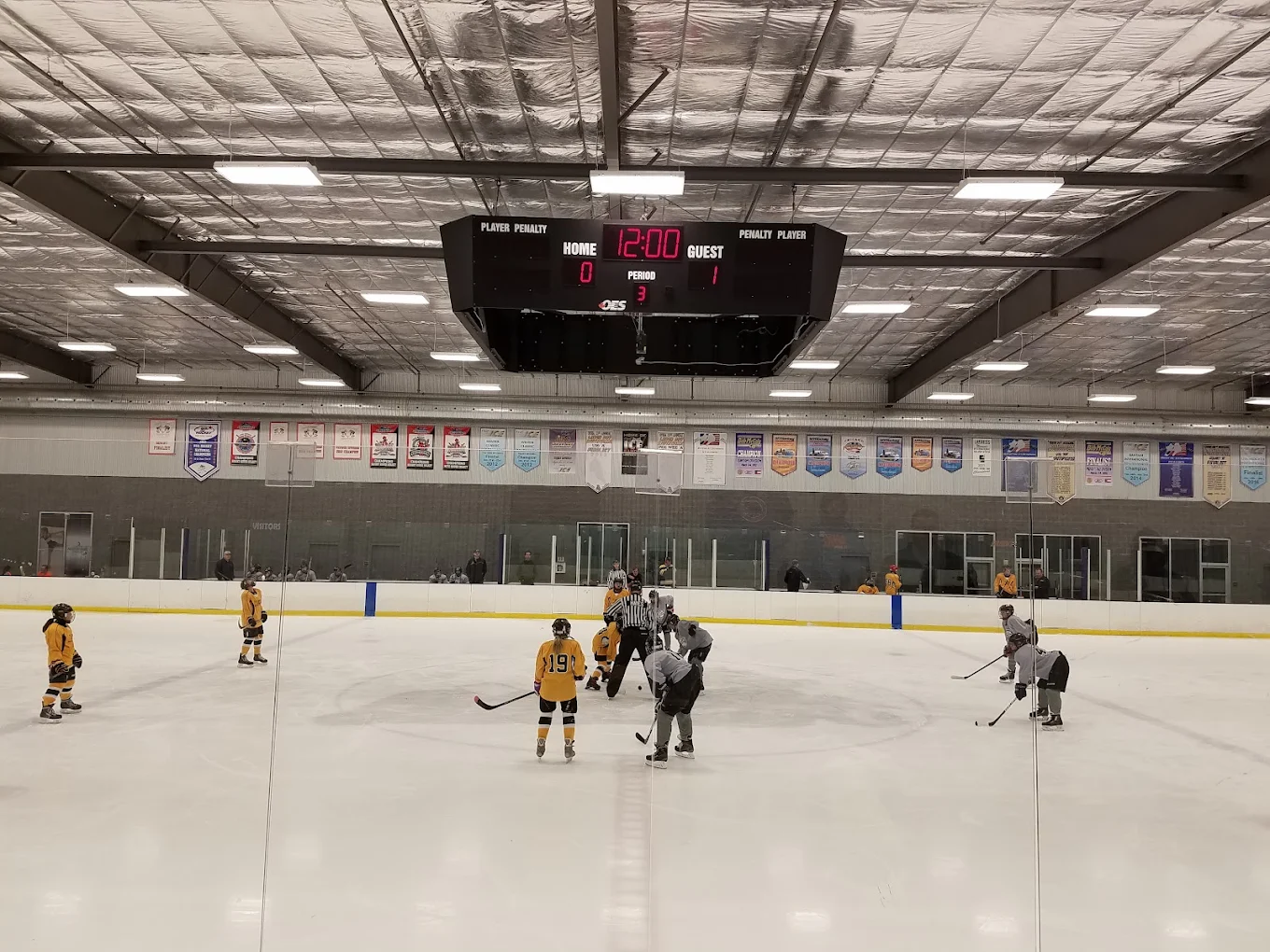 Youth hockey game in The Ice rink, Cumming, GA, players in yellow and gray jerseys, scoreboard showing 12:00 remaining, emphasizing ice skating and hockey activities.