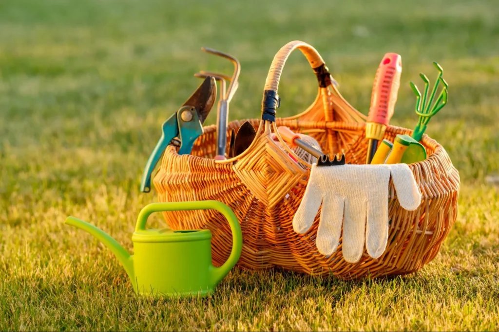 Basket filled with essential lawn care tools including pruning shears, gloves, a watering can, and gardening forks, set against a grassy background.