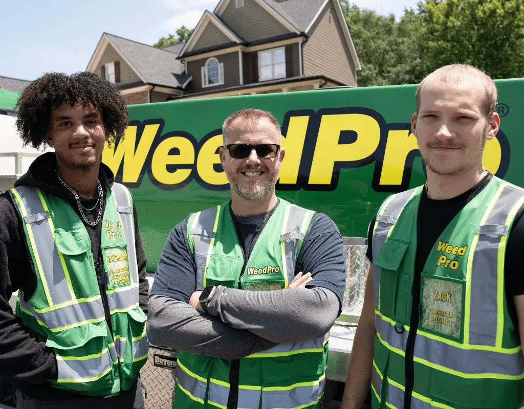 Three Weed Pro Lawn Care team members in green vests, smiling in front of a branded vehicle, showcasing local expertise in irrigation services for Cumming, GA.