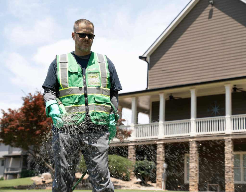WeedPro technician in a green vest spraying a lawn with a handheld wand in front of a large home, demonstrating liquid aeration application.