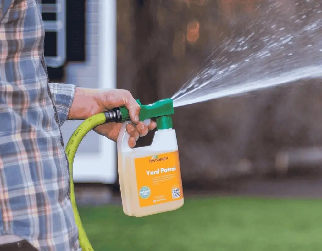 Close-up of a technician’s hand operating a hose-end sprayer dispersing mosquito control solution over a lawn and landscape, creating an insect-free barrier.