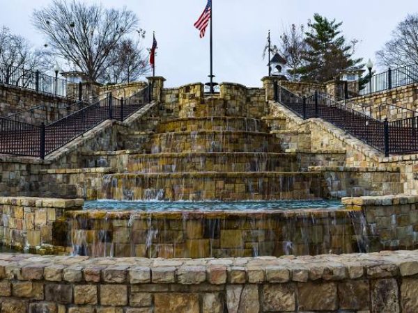 Stone waterfall feature with cascading water, surrounded by stone steps and fences, featuring a flagpole with an American flag in a public space in Norcross, GA.