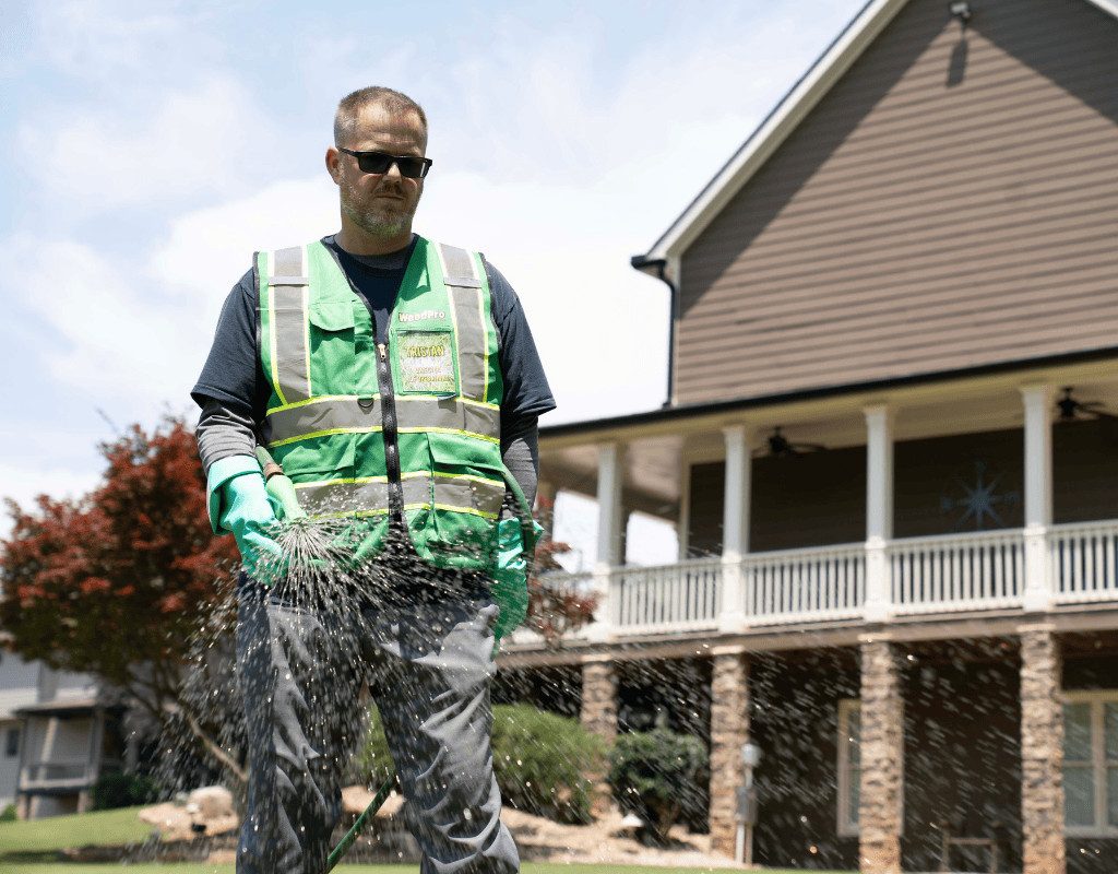 WeedPro technician in protective vest and gloves adjusting a spray wand on a homeowner’s lawn to ensure proper sprinkler performance.