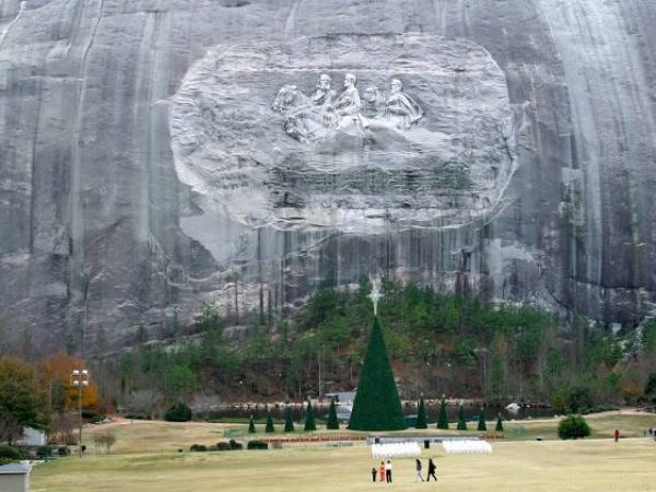 Stone Mountain, GA landscape featuring the iconic Stone Mountain carving of Confederate leaders, surrounded by a green park area with a Christmas tree and visitors walking on the lawn.