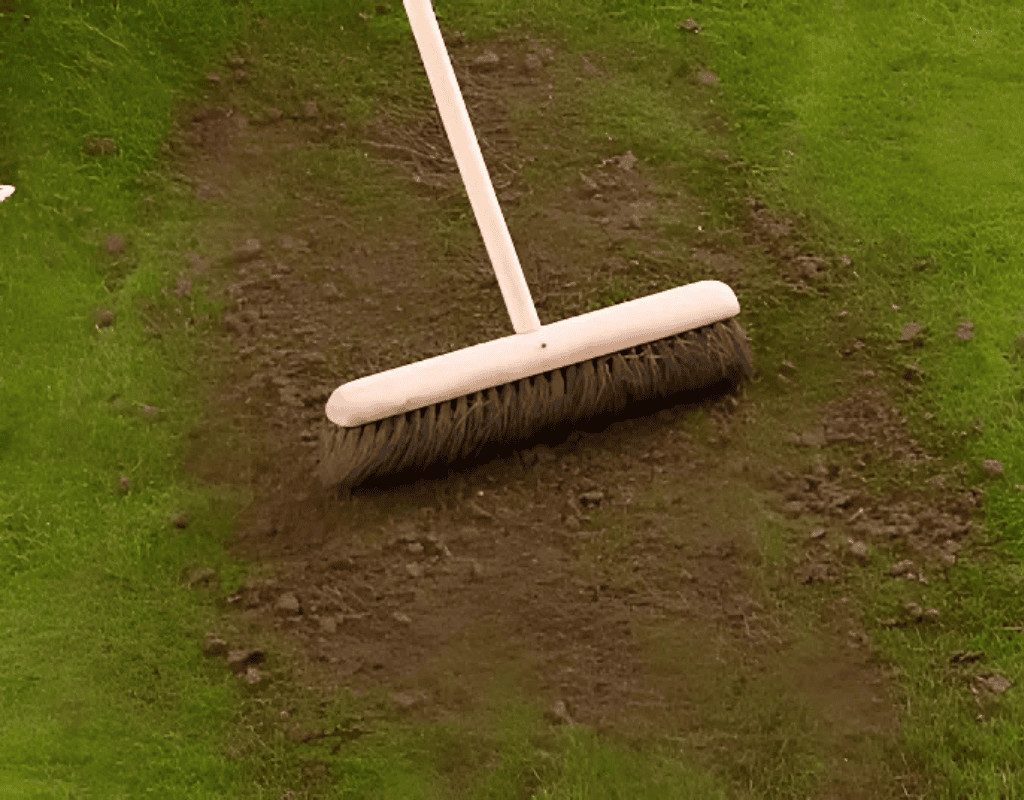 Close view of a push broom smoothing dark top-dressing soil into patches on a green lawn, finalizing the leveling and soil enrichment process.