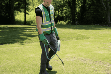 Technician wearing a WeedPro vest and gloves, using a pump sprayer to apply weed control solution across a lawn.